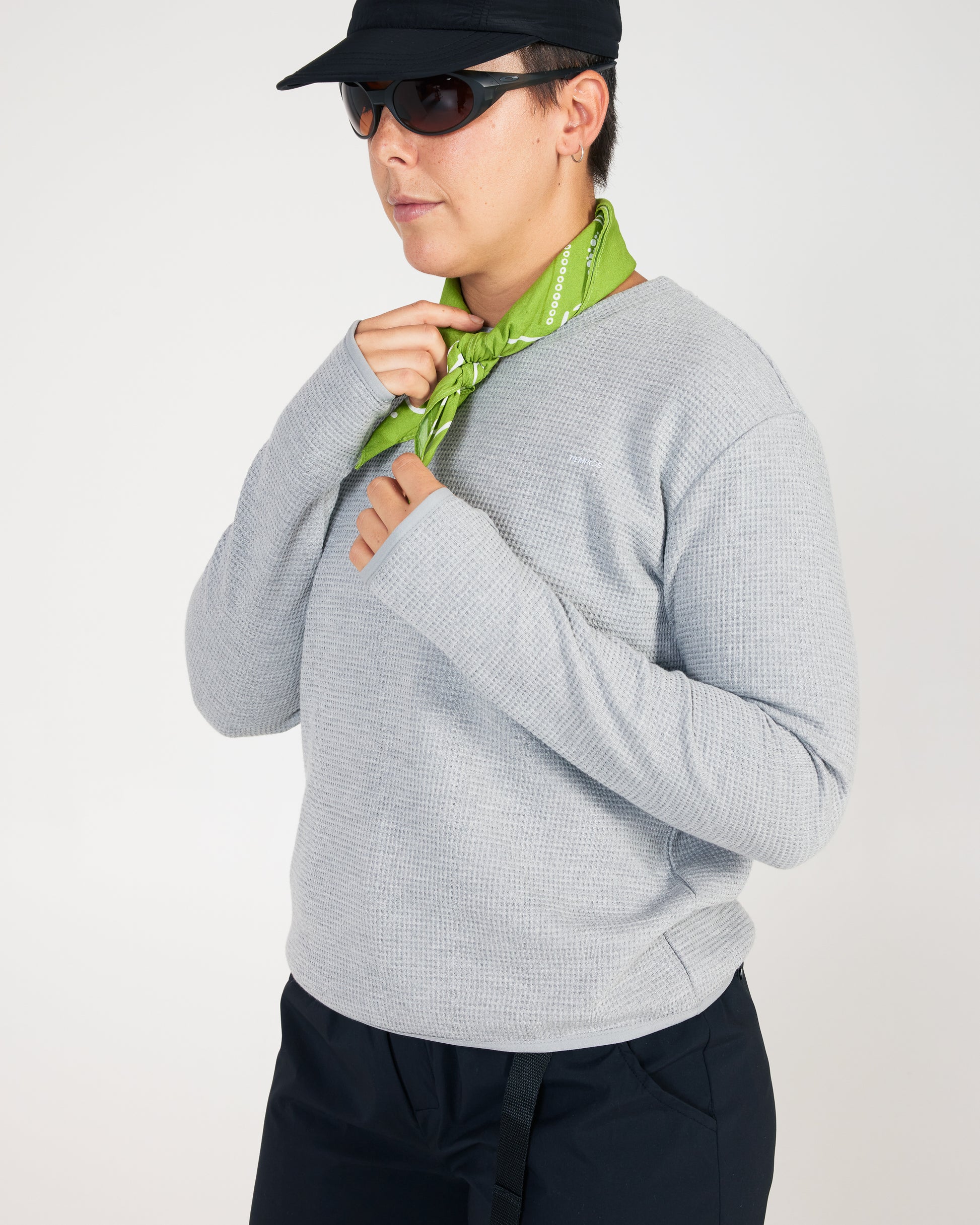 Person wearing a gray sweater, black cap, sunglasses, and a green scarf on a white background